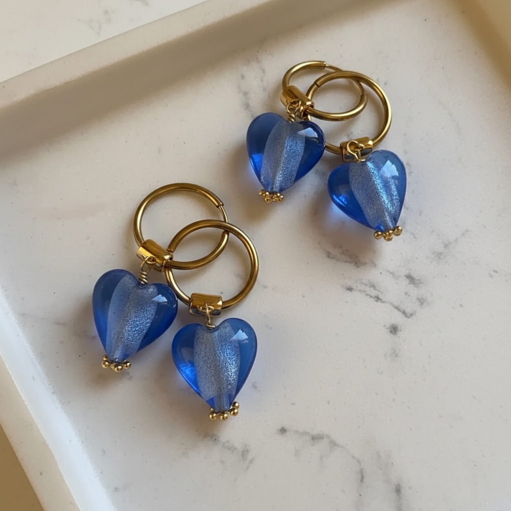 Blue heart-shaped glass earrings with gold loops on a marble surface