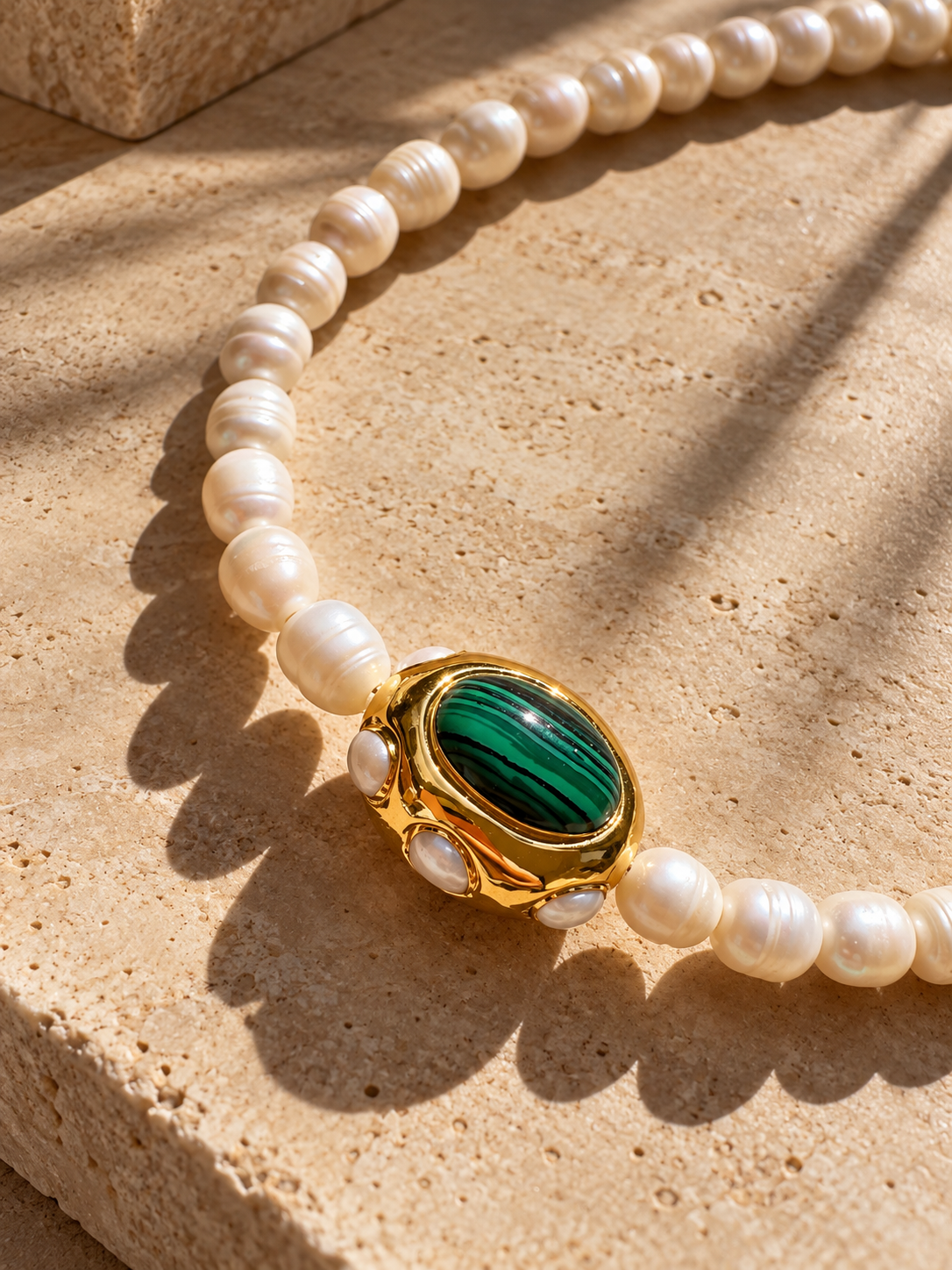 A close-up of AIGLI freshwater pearl and green malachite necklace, resting on a textured beige stone surface.