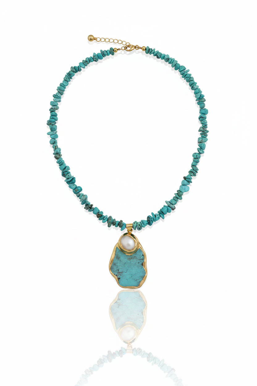 A turquoise beaded necklace with a large raw turquoise stone pendant in a gold frame, featuring a small white pearl accent at the top of the pendant, displayed on a reflective white surface.