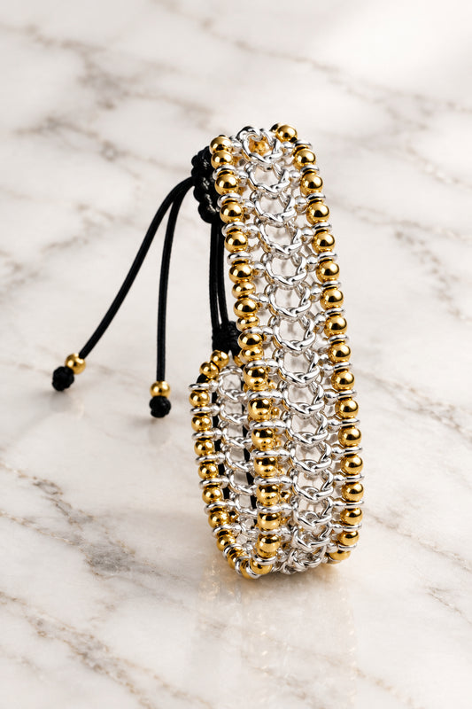 A beaded wrap bracelet with silver and gold metallic beads and black cord ties, displayed on a white marble surface.