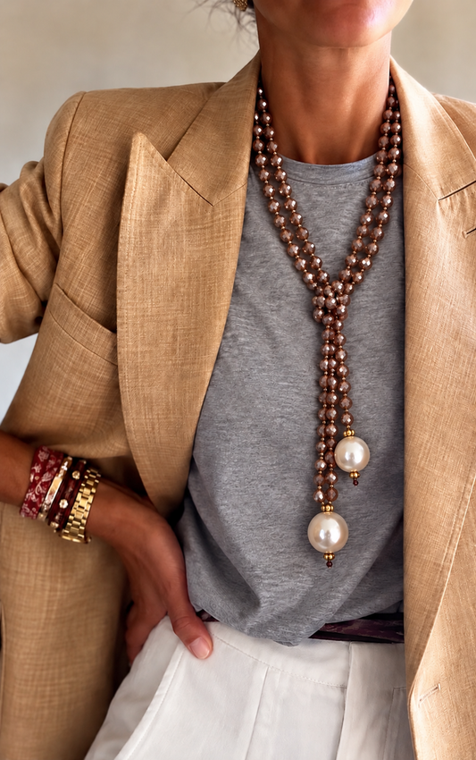 A close-up of a woman wearing a long multi-strand nude beaded lariat necklace with two large white pearl-like drops, styled over a gray t-shirt and tan blazer.