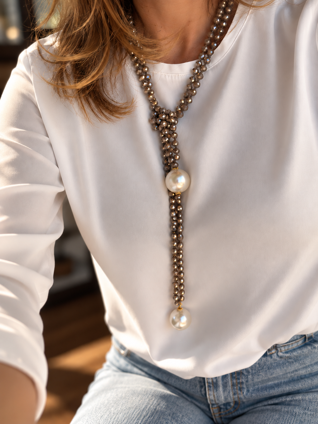 A close-up of a woman wearing a long lariat-style necklace with metallic bronze beads, two large white pearl-like beads, and light blue jeans against a softly blurred indoor background.