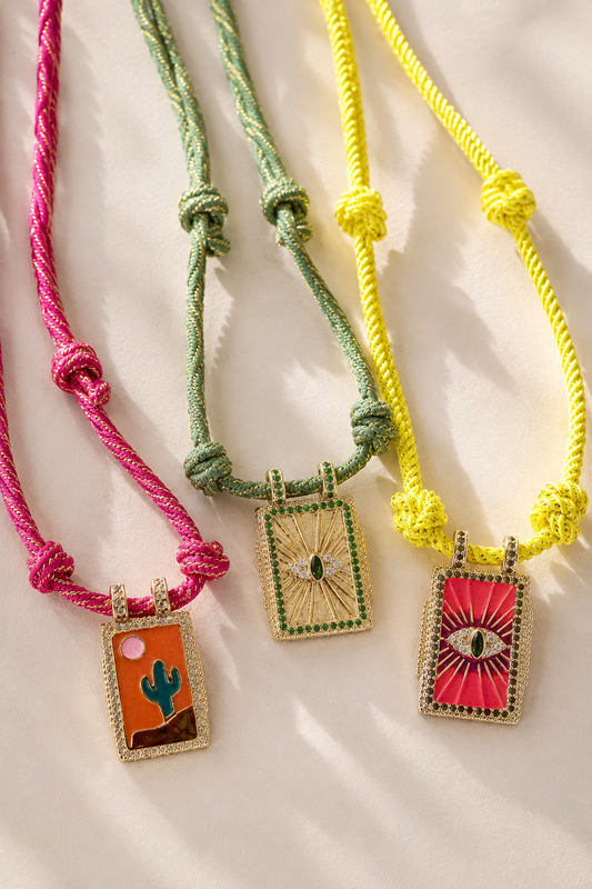Three bohemian-style rope necklaces with rectangular embroidered pendants are arranged diagonally on a light beige background. The necklaces feature pink, sage green, and bright yellow cords, with pendants depicting a desert cactus, a sunburst with a green teardrop, and an eye of providence design.