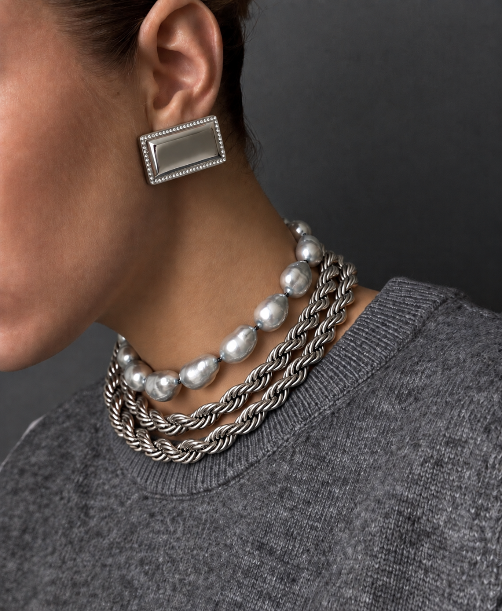 A close-up of a model wearing a layered silver jewelry set, featuring a rectangular silver earring with a beaded border and a multi-strand silver necklace combining twisted rope chains and large baroque pearls, paired with a gray knit sweater against a dark gray background.
