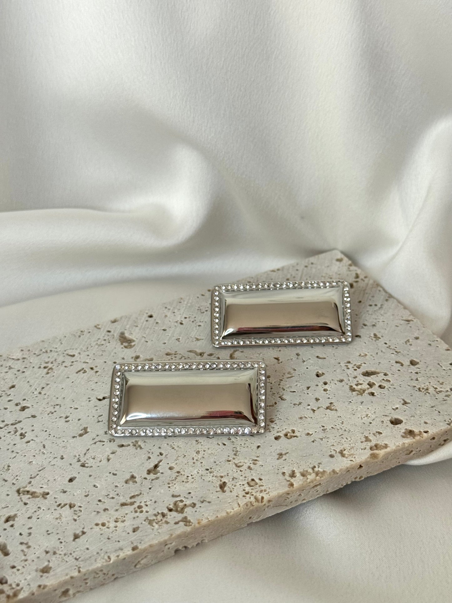 Pair of silver rectangular earrings on a textured surface with white fabric background
