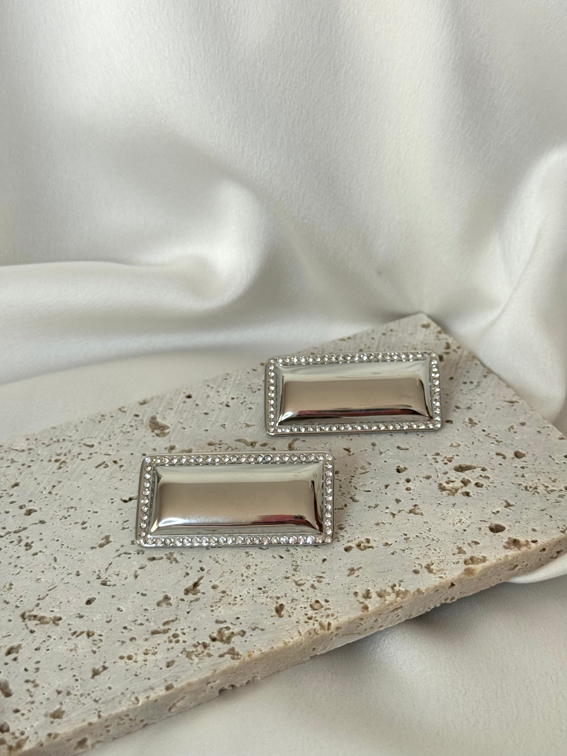 Pair of silver rectangular earrings on a textured surface with white fabric background