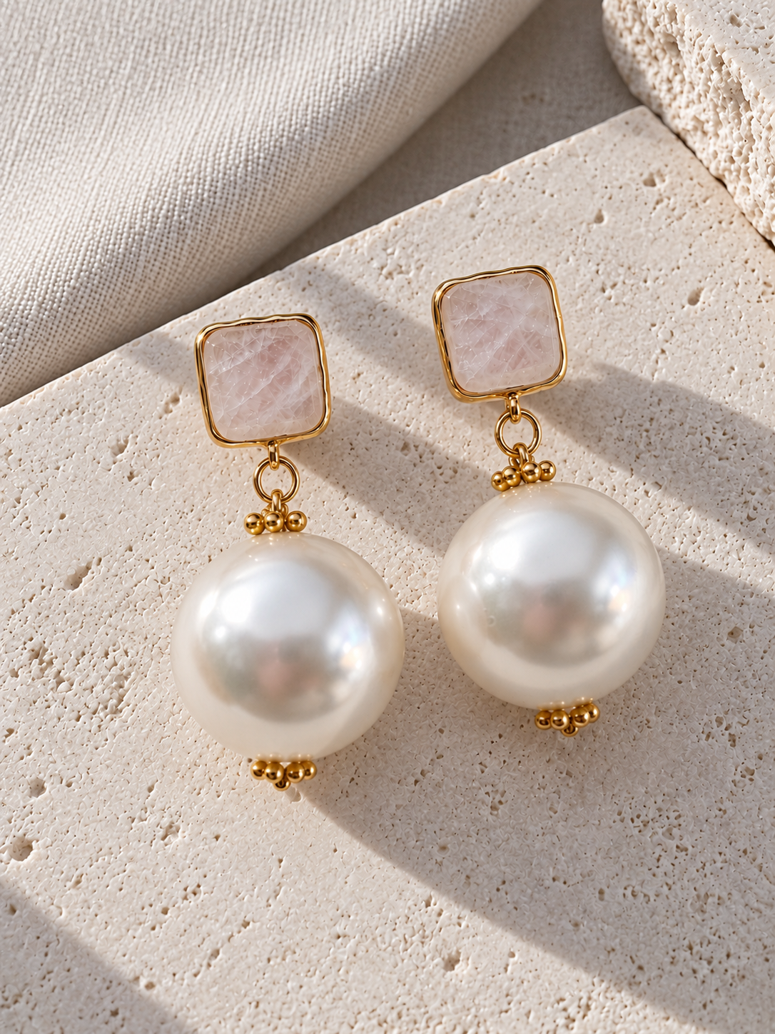 A pair of gold drop earrings featuring square pale pink stone tops and large round white pearls, displayed on a textured beige stone surface.