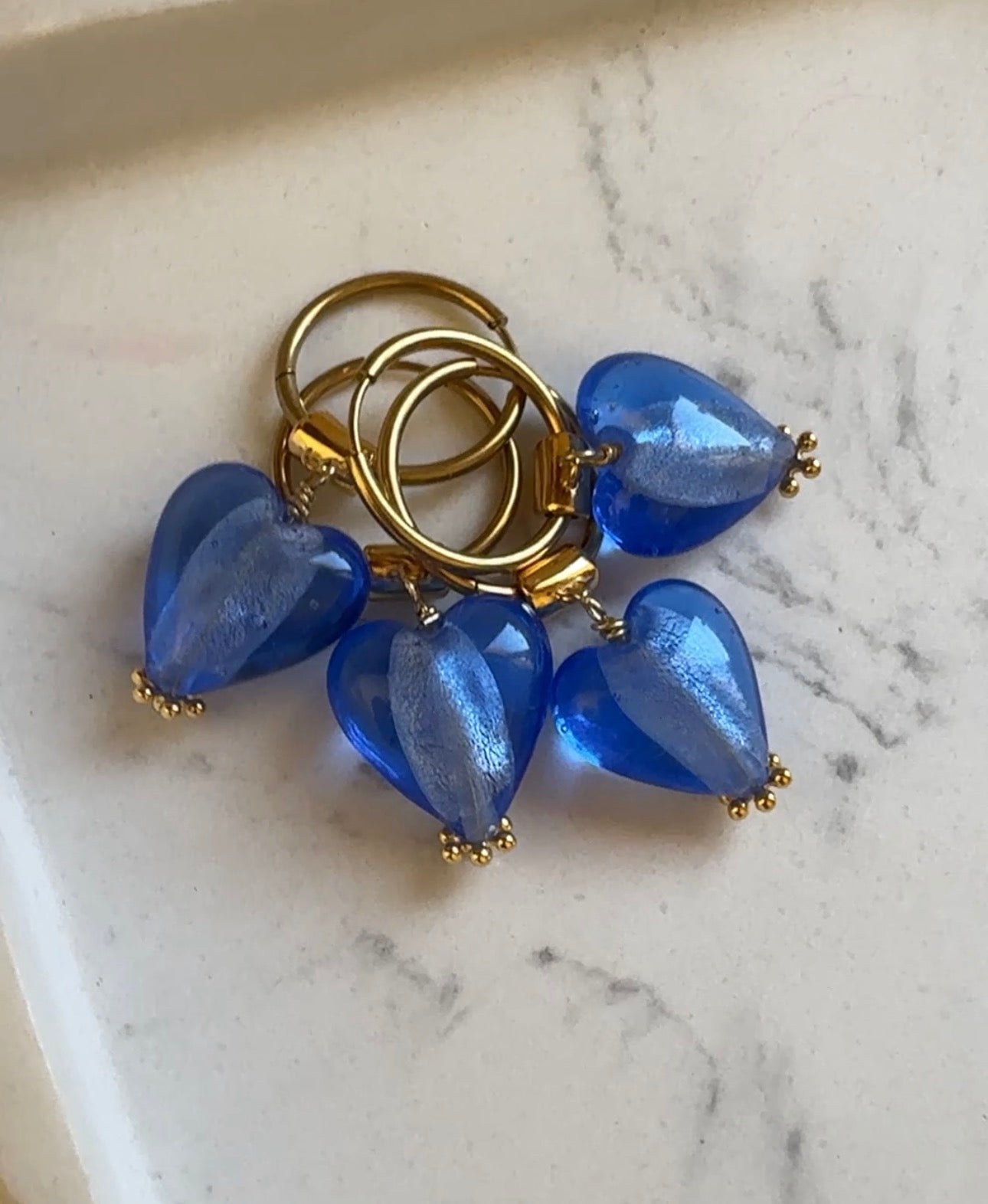 Gold earrings with blue heart-shaped Murano glass beads on a white marble surface