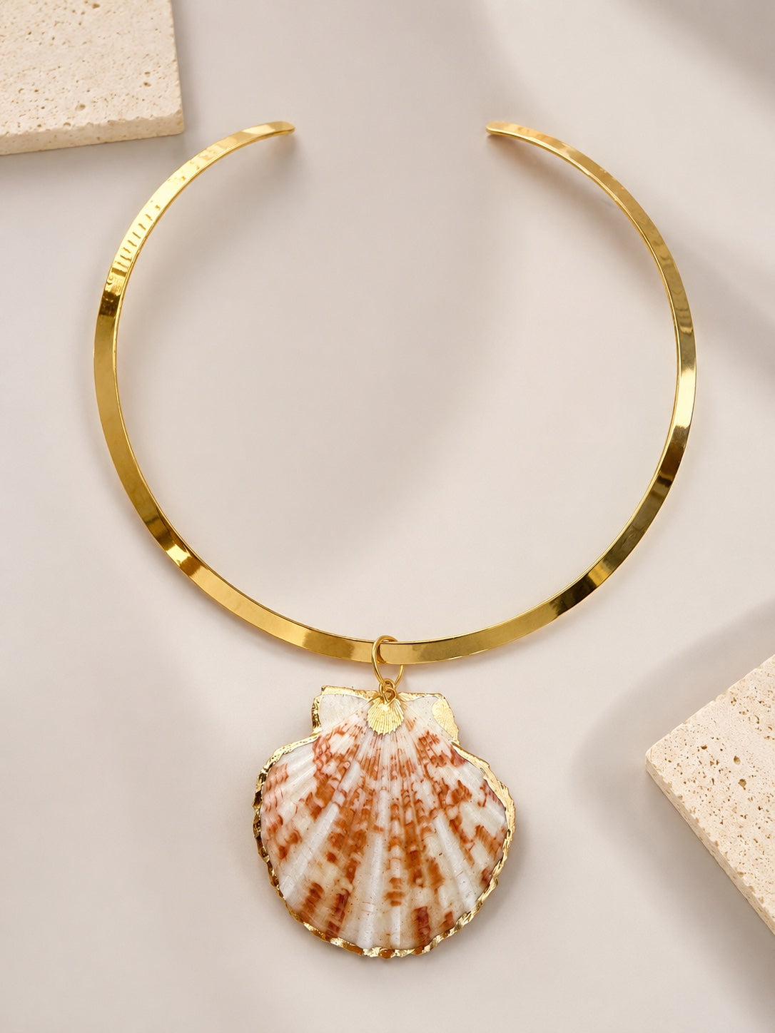 Handmade gold-tone metal collar necklace with a dangling seashell pendant, displayed on a light beige surface with decorative stone tiles by LOTUS by M.  