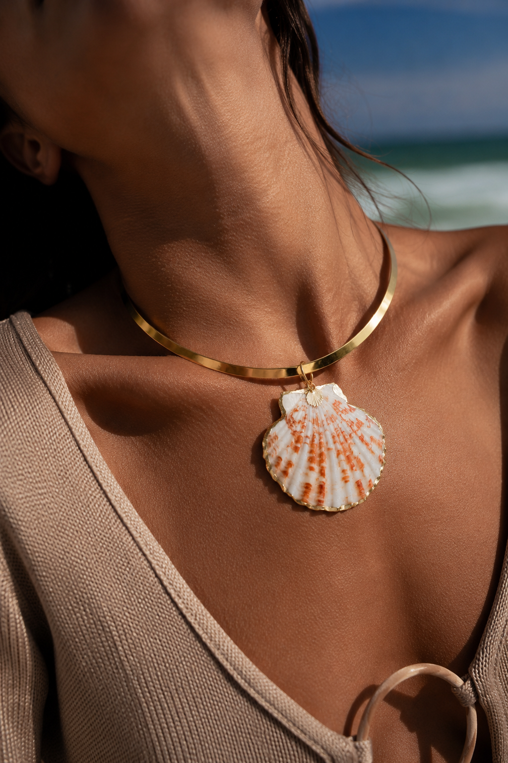 A close-up lifestyle shot of a model wearing a gold-toned metal collar necklace with a large, realistic white and orange seashell pendant, set against a blurred beach background.
