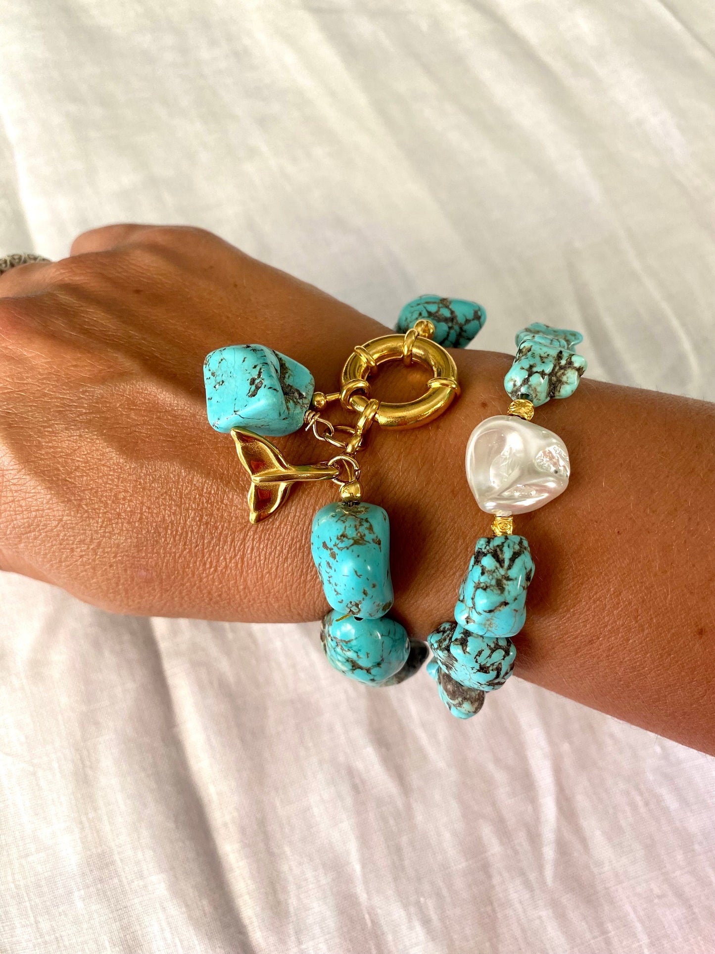 A person's wrist wearing two bracelets; one with irregular aquamarine stones and a gold-plated stainless steel charm, and the other with a large round mother of pearl stone.